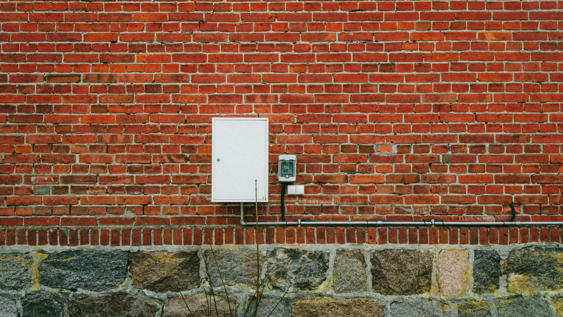 Red brick house wall above a stone base, with a white box fixed to it